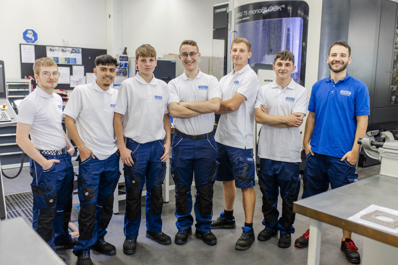 Das Bild zeigt eine Gruppe von sechs lächelnden gewerblichen Azubis in weißen Poloshirts mit Firmenlogo und blauer Arbeitshose, die selbstbewusst mit den Händen in den Taschen oder verschränkten Armen in einer hellen Werkstatt mit ihrem Ausbilder vor einer CNC-Maschine und einem Maschinenbedienfeld posieren.