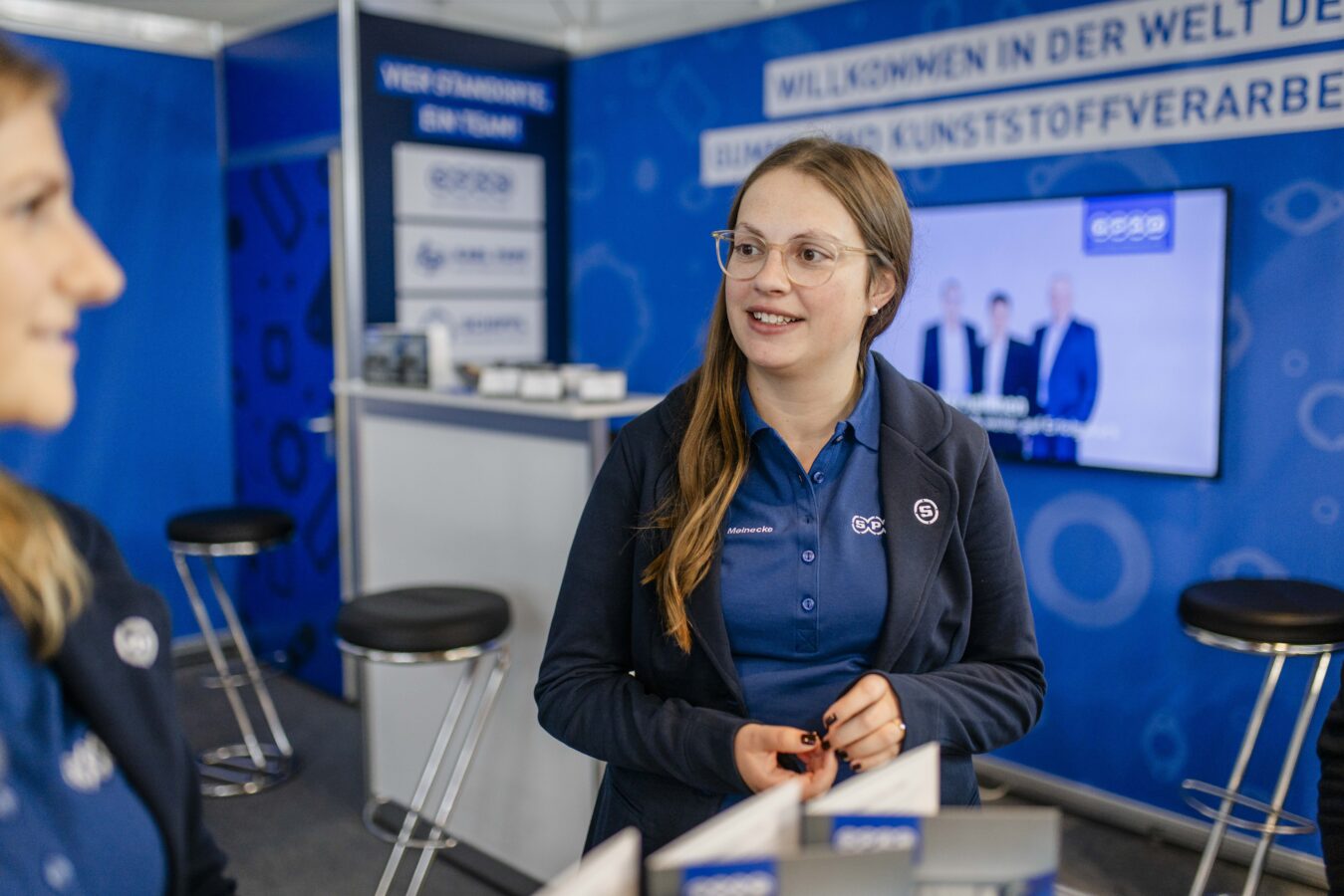 Eine lächelnde Mitarbeiterin im blauen Team-Outfit führt ein Gespräch an einem Messestand vor einem blauen Hintergrund mit der Aufschrift „Willkommen in der Welt der Kunststoffverarbeitung“.