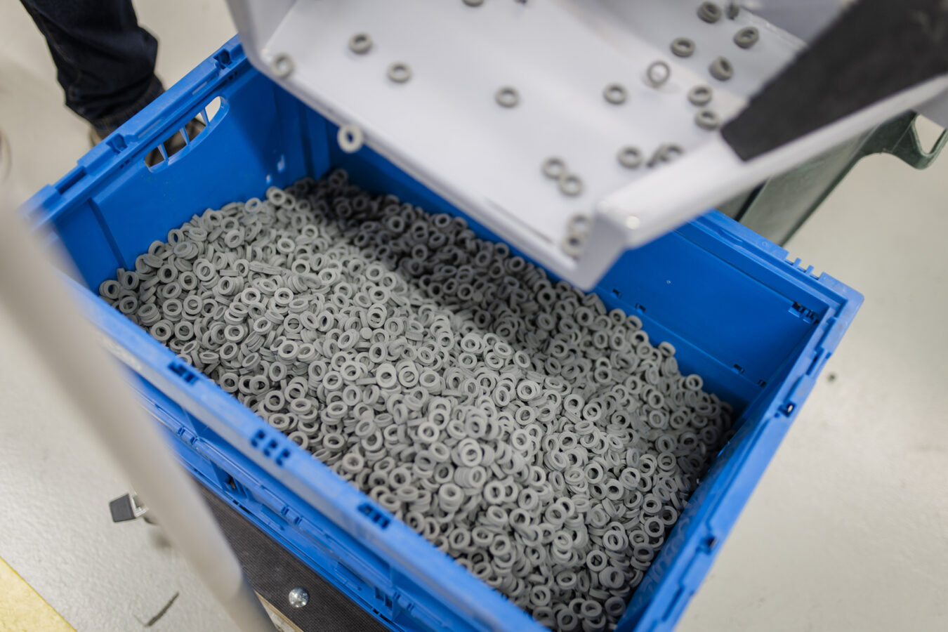 Eine Nahaufnahme zeigt eine blaue Späh-Kiste, die mit einer großen Menge kleiner, grauer Dichtungsringe gefüllt ist, während weitere Ringe aus einem darüber befindlichen weißen Behälter hineingeschüttet werden