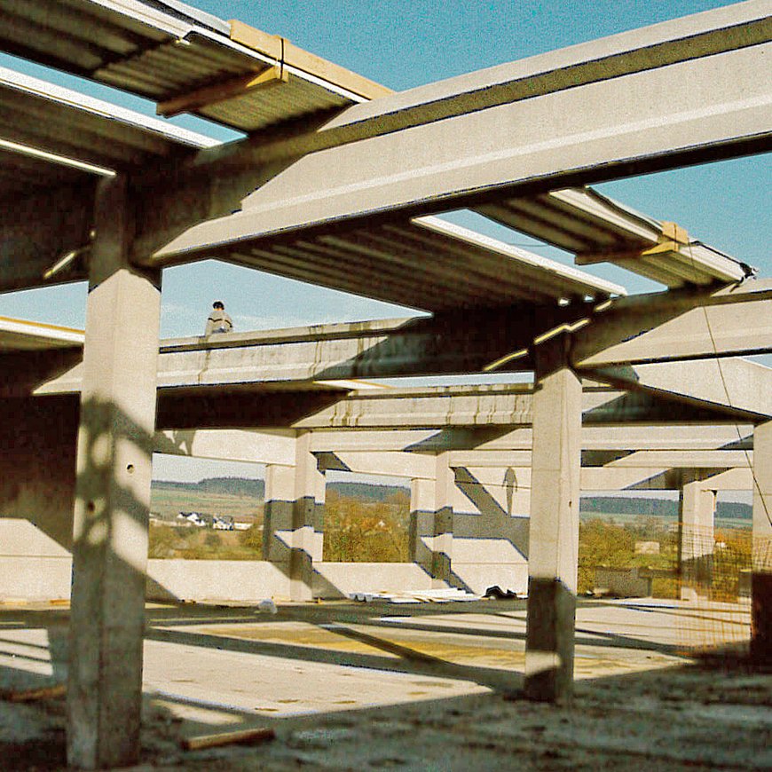 Die Baustelle eines Gewerbegebäudes im Jahr 1993 zeigt eine Skelettkonstruktion aus massiven Betonpfeilern und -trägern, mit einem Arbeiter, der auf einem Träger steht, und einer dahinterliegenden Landschaft