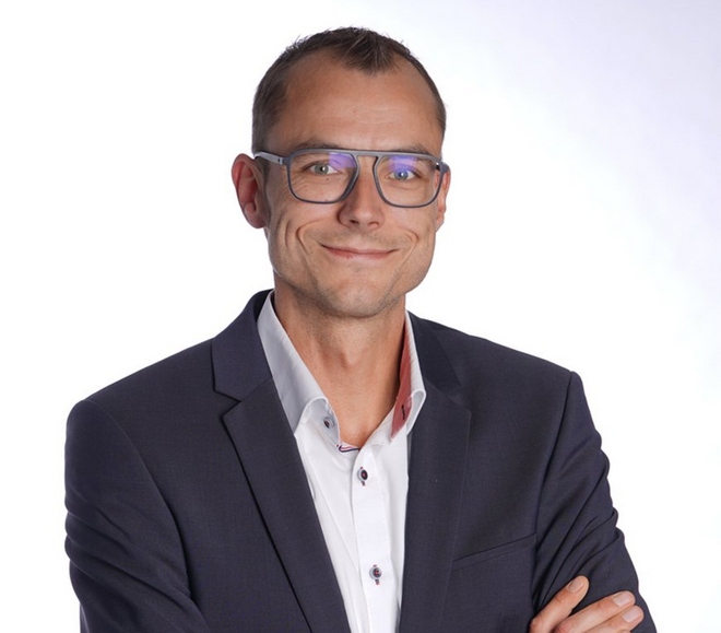 Das professionelle Porträt zeigt Thomas Späh, einen lächelnden Mann mit graublauer, rechteckiger Brille, in einem dunkelblauen Anzug über einem weißen Hemd mit geöffnetem Kragen.