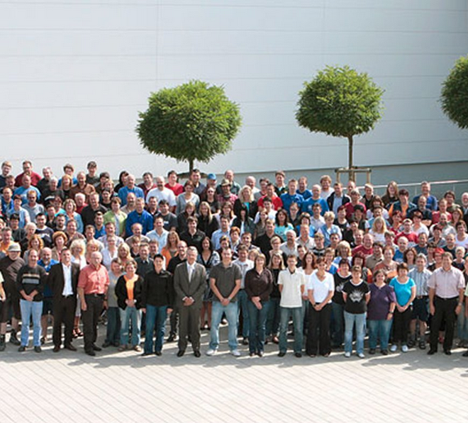 Das Bild zeigt eine große Gruppe von Mitarbeitern der Firma Späh, die vor einer hellen Gebäudefront und zwischen zwei runden Bäumen für ein Gruppenfoto im Freien posieren.