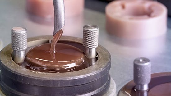 Eine Nahaufnahme zeigt, wie eine braune, zähflüssige Masse in eine Metallform gegossen wird.