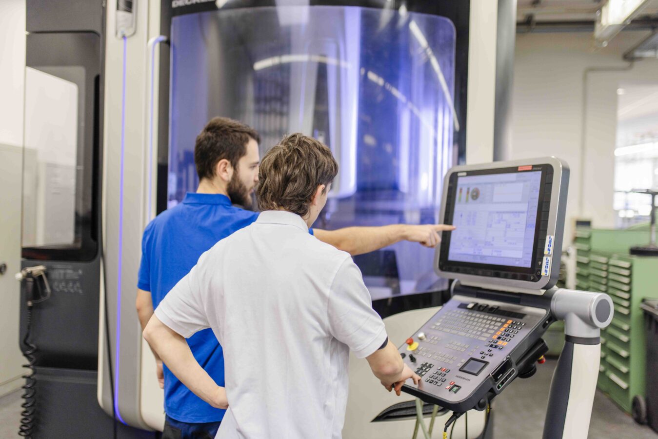 Das Bild zeigt den Ausbilder und einen Azubi, die gemeinsam konzentriert vor einem großen, modernen, graublauen Maschinengehäuse stehen, wobei der Azubi an der Steuerung arbeitet und der Ausbilder ihm etwas auf dem Steuerungsdisplay zeigt.