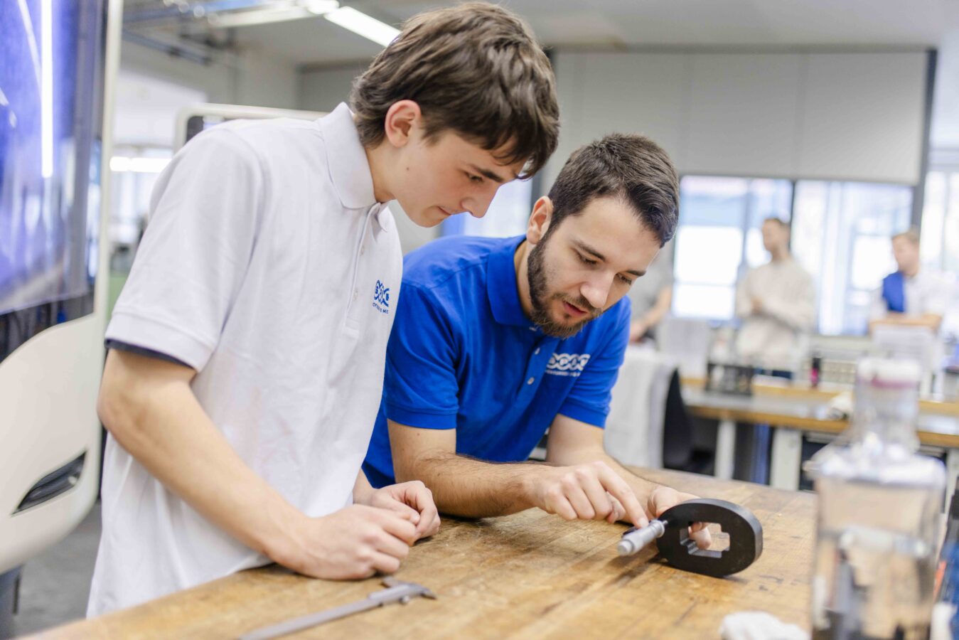 Das Bild zeigt einen Ausbilder und einen Azubi in einer Werkstatt, die sich über einen hölzernen Tisch beugen und gemeinsam ein schwarzes, komplex geformtes Werkstück prüfen.