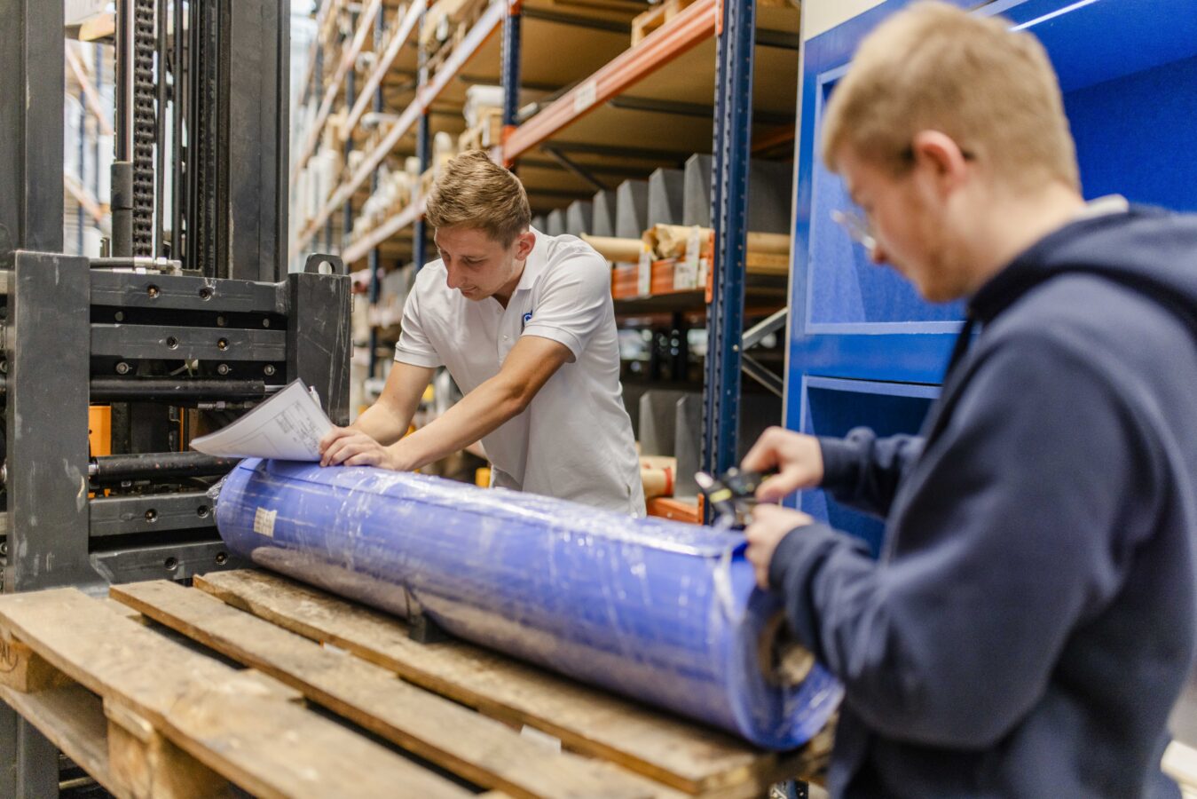 Das Bild zeigt zwei Azubis im Lager: Der linke im weißen Poloshirt notiert etwas auf einem Dokument, das auf einer großen blauen Materialrolle liegt, während der rechte mit einem Messwerkzeug die Rolle ausmisst, die auf einer Palette auf einem Gabelstapler liegt.