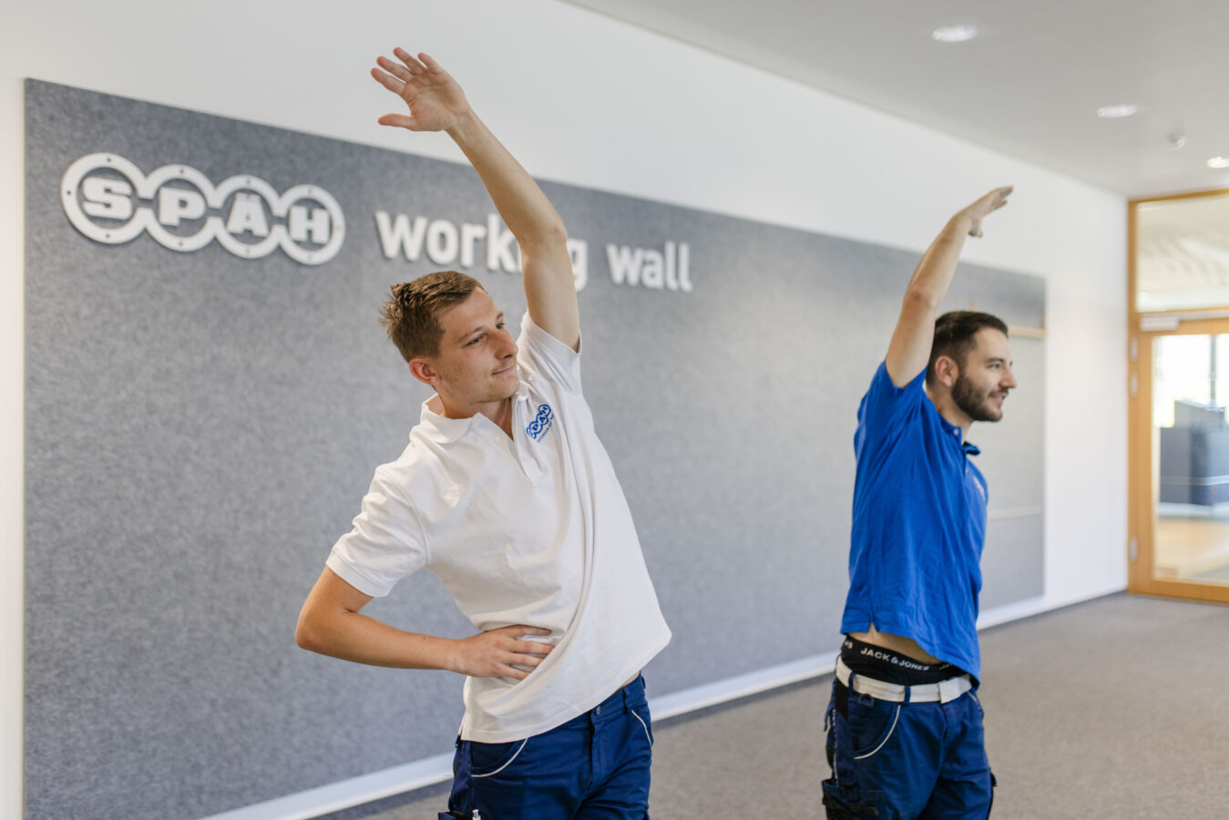 Das Bild zeigt zwei lächelnde Späh-Mitarbeiter, die vor einer grauen Wand mit der Aufschrift 