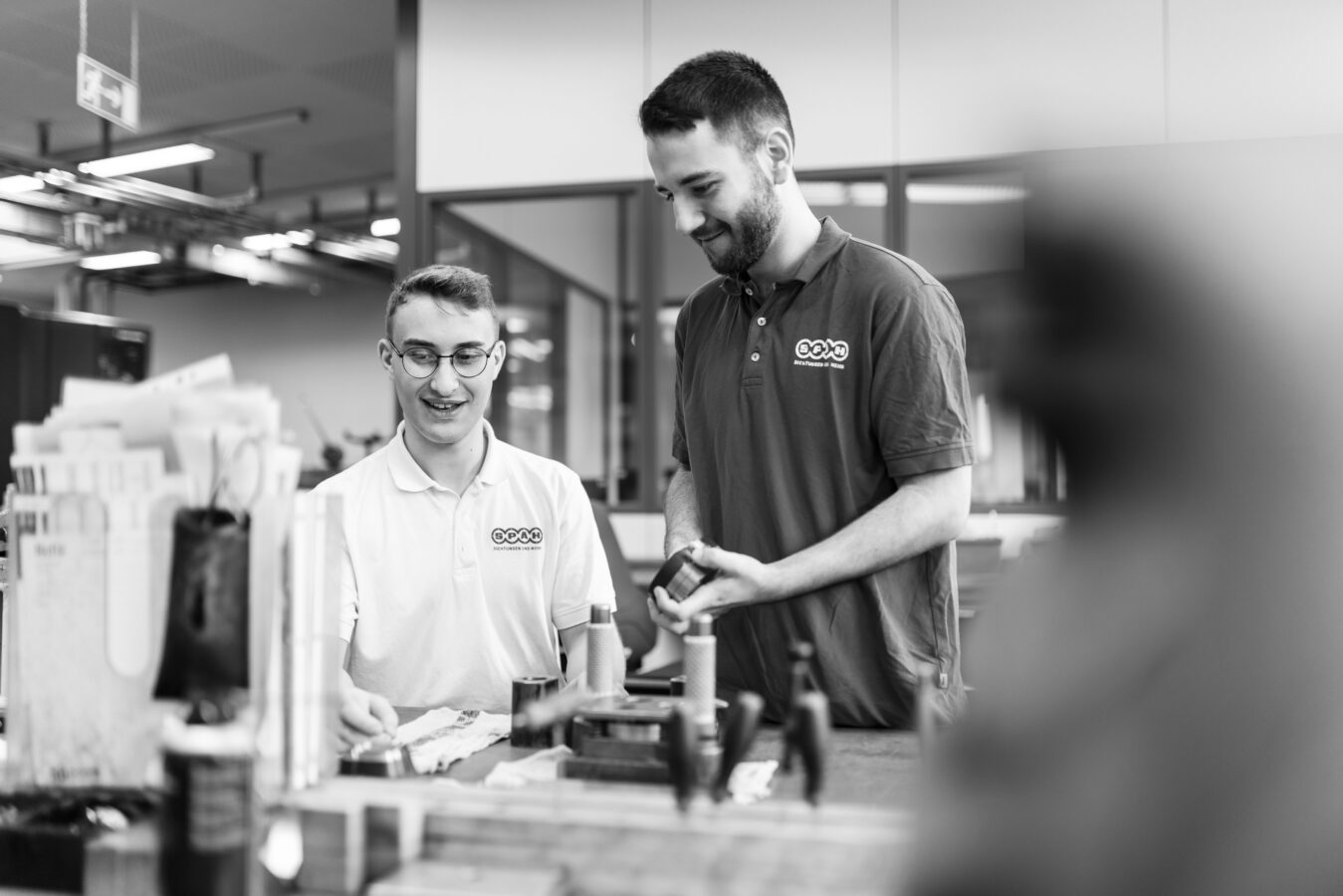 Das Schwarz-Weiß-Bild zeigt den lächelnden Azubi und den lächelnden Ausbilder in einer Werkstatt, die gemeinsam über einem Tisch stehen und ein kleines, dunkles Bauteil in ihren Händen betrachten.