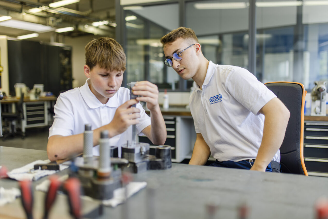 Das Bild zeigt zwei Azubis in weißen Poloshirts in einer Werkstatt, von denen einer konzentriert an einem komplexen, dunklen Bauteil arbeitet und es mit einem silbernen Werkzeug prüft, während der andere ihm aufmerksam zusieht.