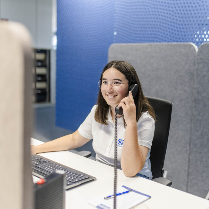 Das Bild zeigt eine lächelnde Azubine im weißen Poloshirt im Büro, die konzentriert und freundlich ein Telefongespräch an einem hellen Schreibtisch vor einer blauen und grauen Trennwand führt.