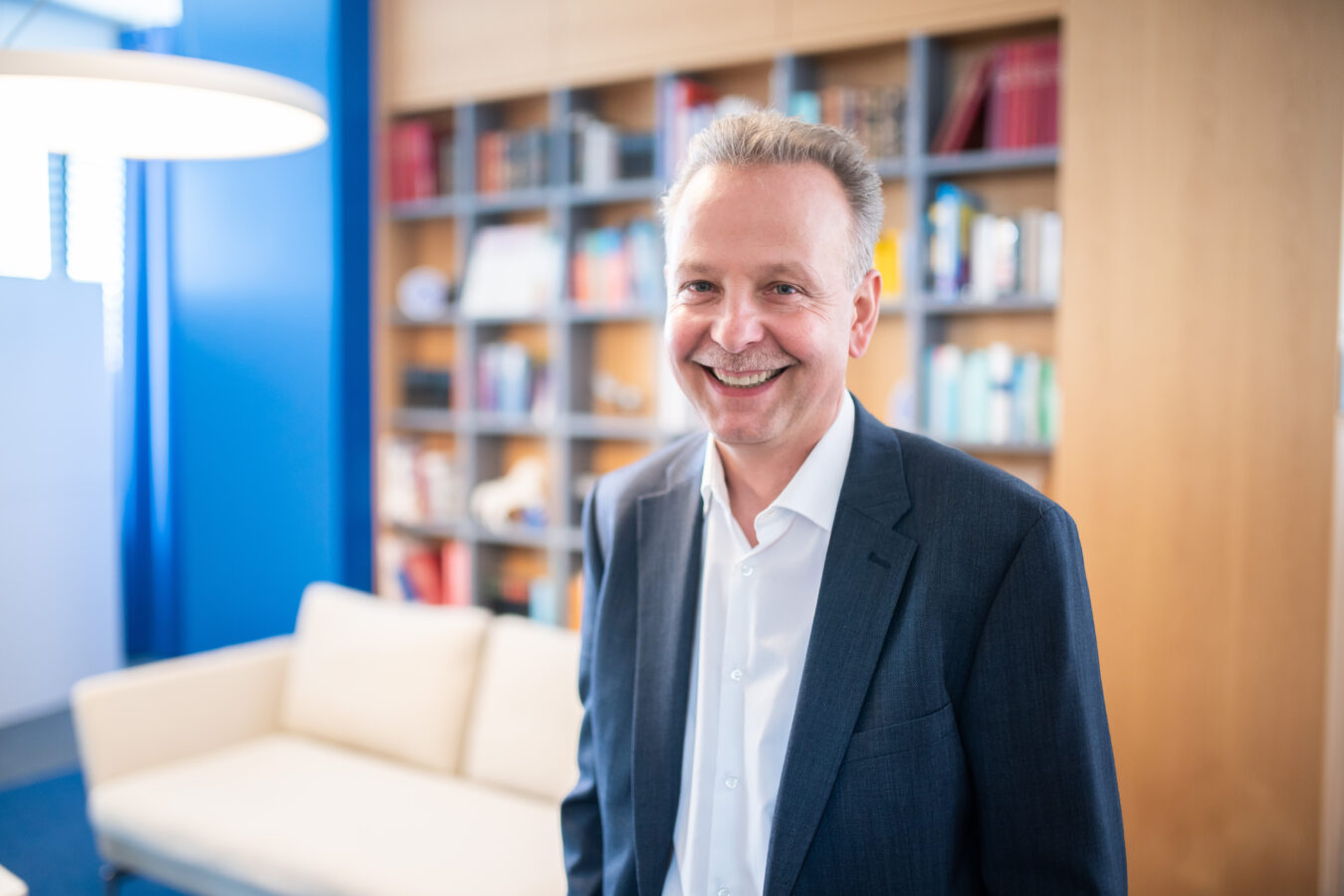 Ein lächelnder, mittelalter Mann in einem dunkelblauen Anzug und einem weißen Hemd steht in einem hellen, modernen Raum vor einem Bücherregal mit Holzrahmen und einem weißen Sofa.