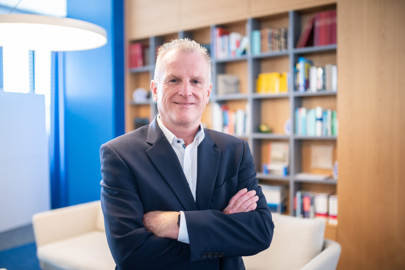 Ein lächelnder, mittelalter Mann in einem dunkelblauen Anzug und einem weißen Hemd steht mit verschränkten Armen vor einem Bücherregal mit Holzrahmen und einem weißen Sofa.