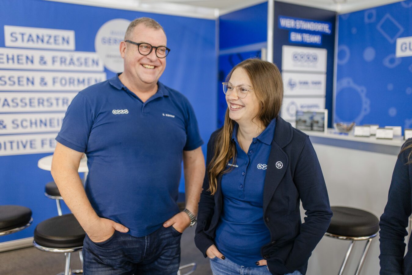 Zwei lächelnde Späh-Mitarbeiter, bekleidet in blauen Poloshirts und Jeans, stehen vor einem blauen Messestand der Firma Späh, der ihre Dienstleistungen wie STANZEN, DREHEN & FRÄSEN und LASERSTRAHL- & WASSERSTRAHL-SCHNEIDEN bewirbt.