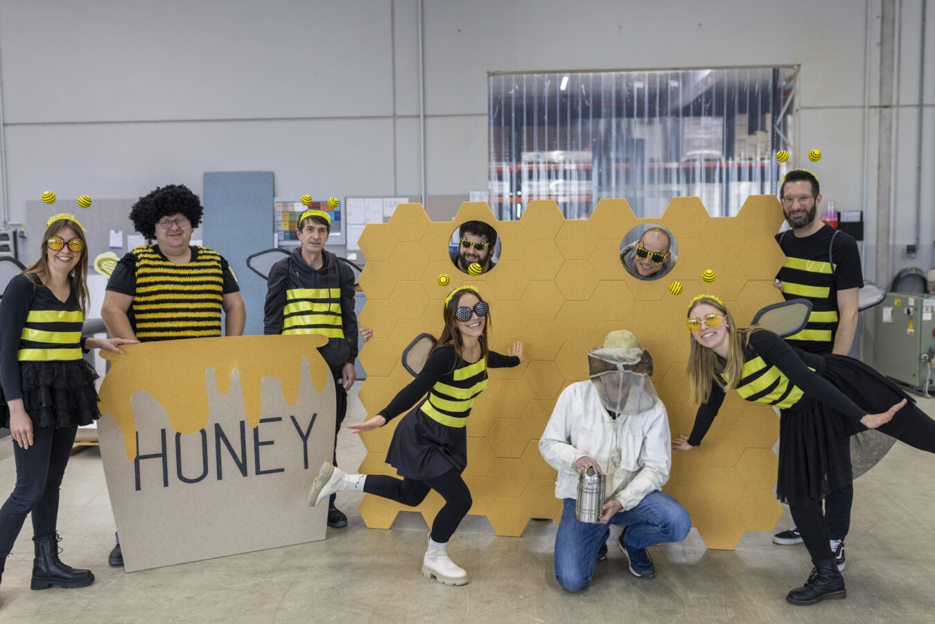 Eine Gruppe von Späh-Mitarbeitern ist in Bienenkostümen und Sonnenbrillen in einer Industriehalle vor einer großen, honiggelben Wabenstruktur aus Pappe und einem Schild mit der Aufschrift "HONEY" versammelt, während ein Mitarbeiter ein Imkerkostüm trägt.