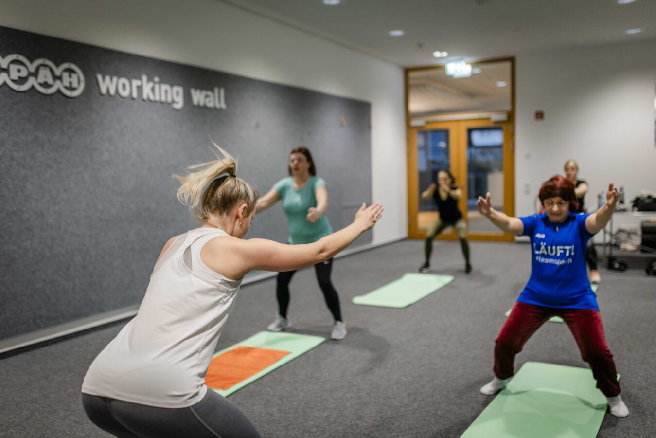 Fünf Späh-Mitarbeiterinnen nehmen an einem Indoor-Workout teil und führen Squats auf Yoga-Matten in einem modernen Raum mit einer grauen Wand, auf der das Späh-Logo und 