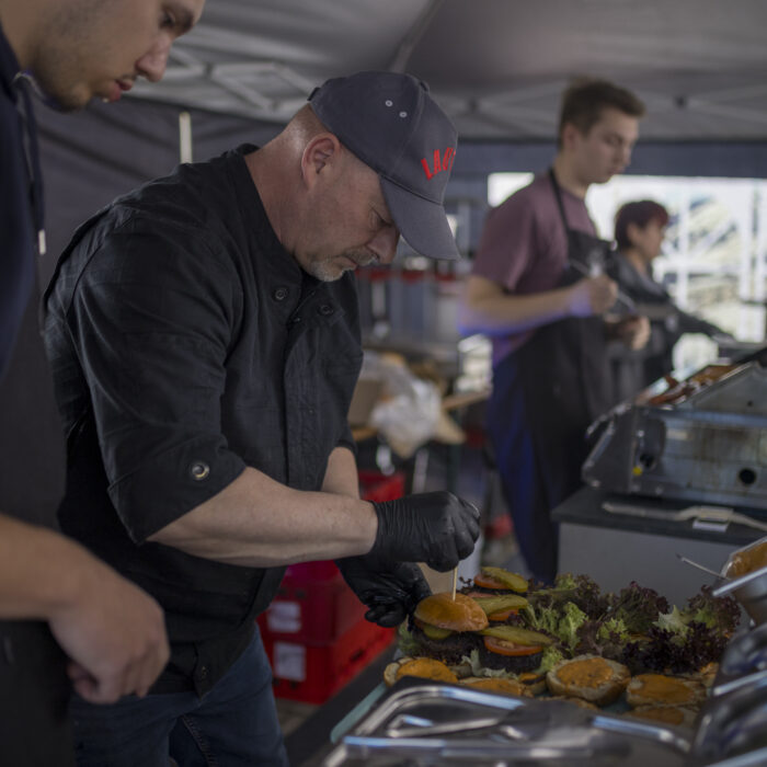 Ein Koch in schwarzer Arbeitskleidung und Kappe belegt beim Grillfest der Firma Späh Burgerbrötchen an einer Theke, während ein junger Mitarbeiter zusieht und weitere Personen im Hintergrund Speisen zubereiten.