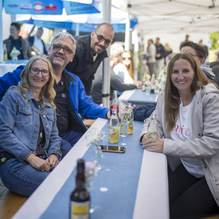 Eine Gruppe von vier Späh-Mitarbeitern sitzt lachend an einem Tisch bei einer Outdoor-Firmenveranstaltung, wobei im Vordergrund zwei Frauen sitzen und im Hintergrund zwei Männer posieren.