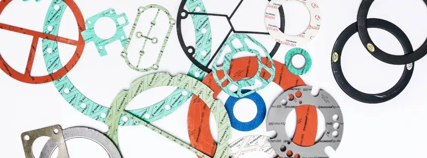 Das Bild zeigt eine Vielzahl von Dichtungen aus verschiedenen Materialien in leuchtenden Farben, darunter Blau, Rot, Türkis, Grün, Schwarz und Weiß, die auf einer weißen Oberfläche verstreut sind.