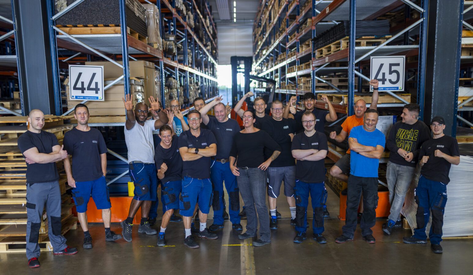 Die Abteilung Lager der Späh-Gruppe posiert als großes Teamfoto inmitten der Hochregale eines Lagers, wobei sie in dunkler Arbeitskleidung stehen, lächeln und zum Teil winken oder die Daumen nach oben halten.