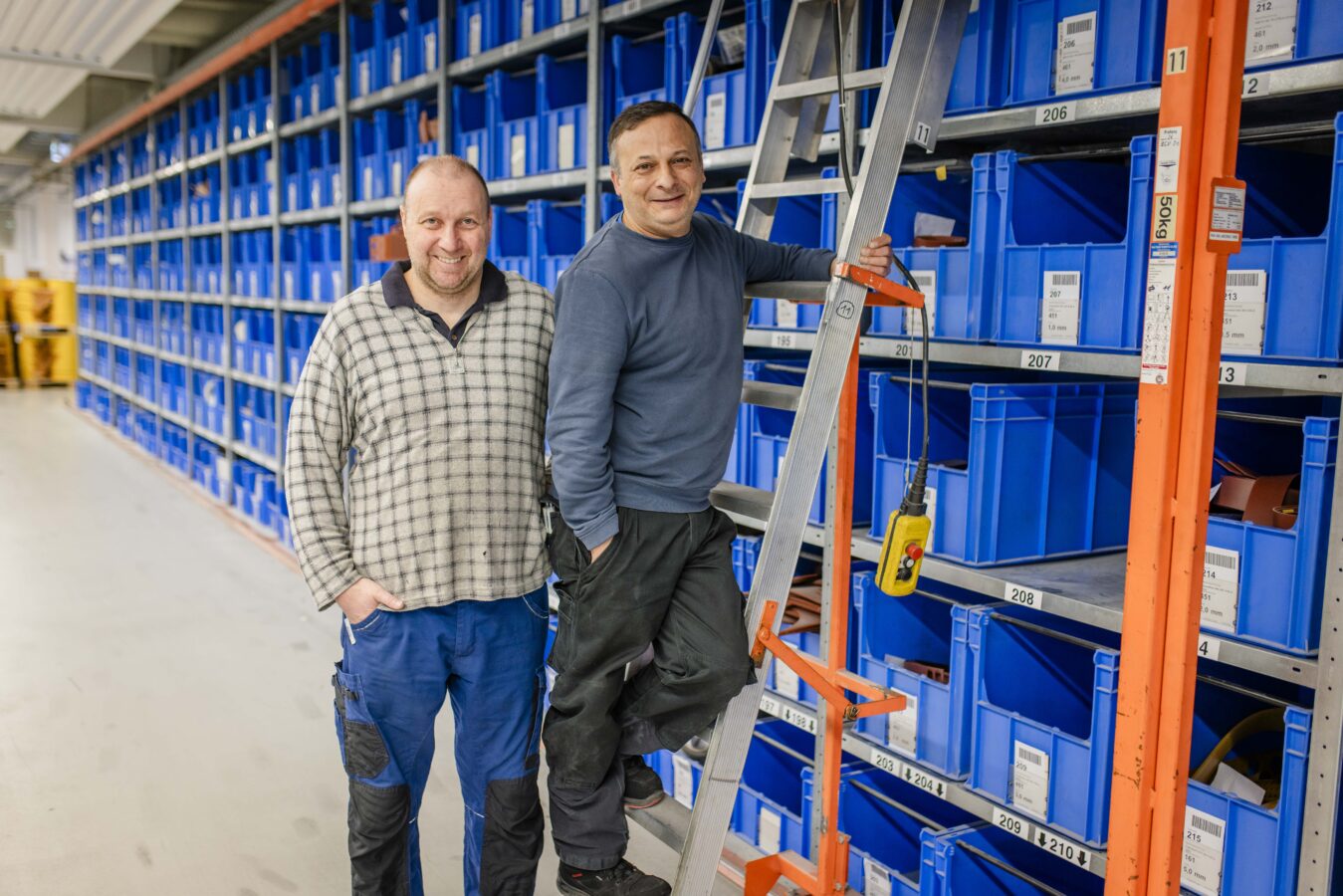 Zwei lächelnde Späh-Mitarbeiter stehen in einem Lagergang mit kleinen blauen Kunststoffkästen: Der linke Mann in einem karierten Hemd steht auf dem Boden, und der rechte Mann in einem blauen Pullover steigt gerade auf einer silbernen Leiter an einem der Regale hoch.