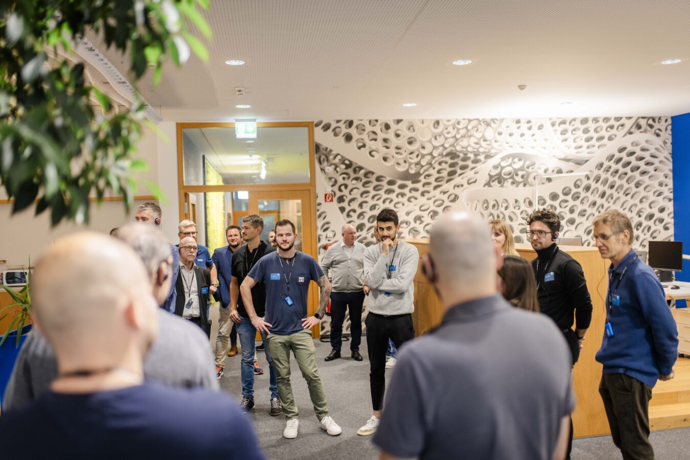 Eine Gruppe von Gästen hört einer jungen Frau in einem schwarzen T-Shirt aufmerksam zu, die vor einer auffälligen, weiß-gemusterten Wand in einem modernen Innenraum steht und eine Führung leitet.