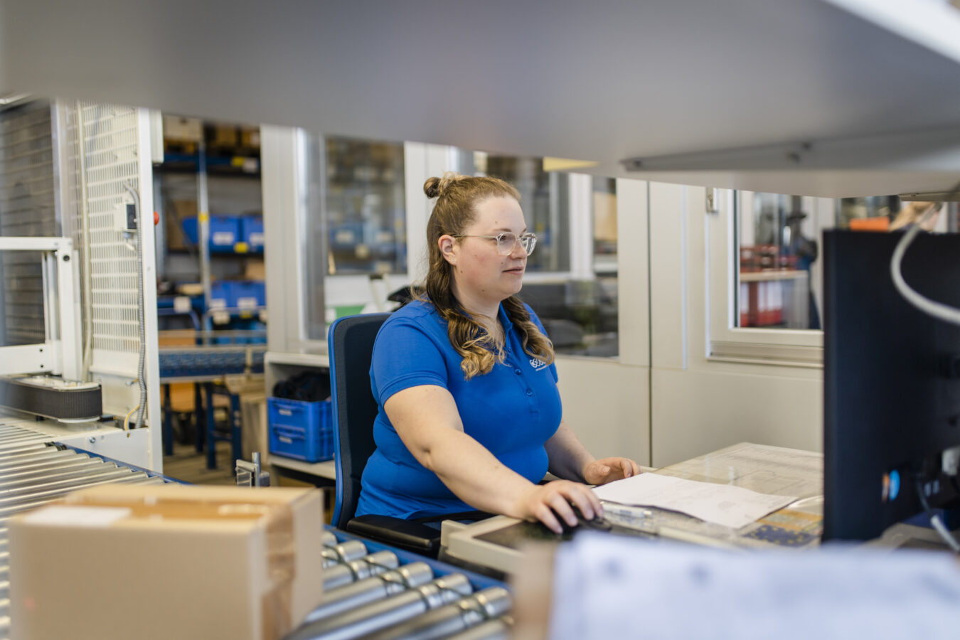 Eine Mitarbeiterin in einem blauen Poloshirt arbeitet konzentriert an einem Computerarbeitsplatz in einer Logistikumgebung.