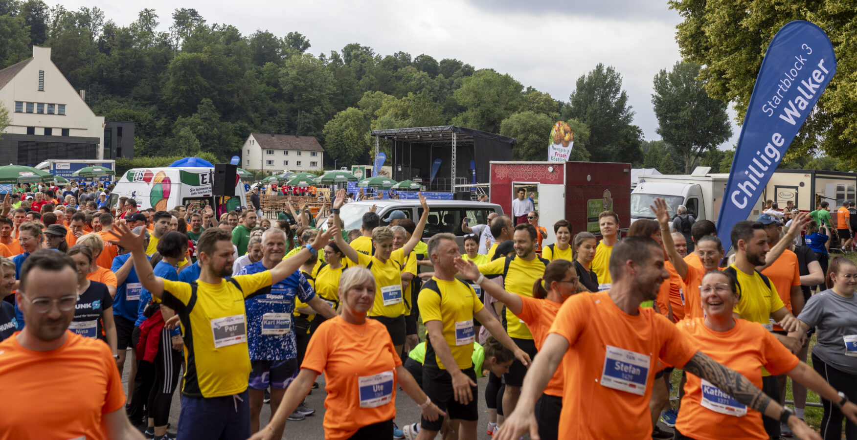 Eine große Gruppe von Läufern und Teilnehmern, viele davon in blauen, gelben oder orangefarbenen T-Shirts, steht mit ausgestreckten Armen, kurz vor dem Start eines Lauf-Events.
