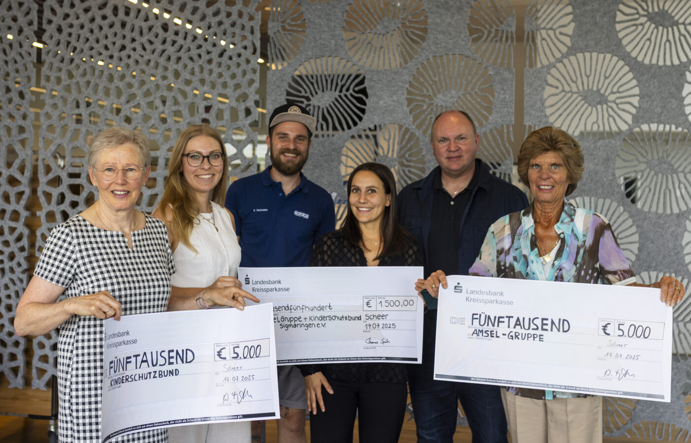 Eine Gruppe von sechs Personen, drei Frauen und drei Männer, hält Schecks der Landesbank Kreissparkasse im Wert von € 5.000 für den Kinderschutzbund und die Amsel-Gruppe sowie einen Scheck über € 1.500 für die Jugend- und Kinderkrebshilfe Sigmaringen in einem modernen Innenraum mit einer gemusterten Trennwand im Hintergrund.