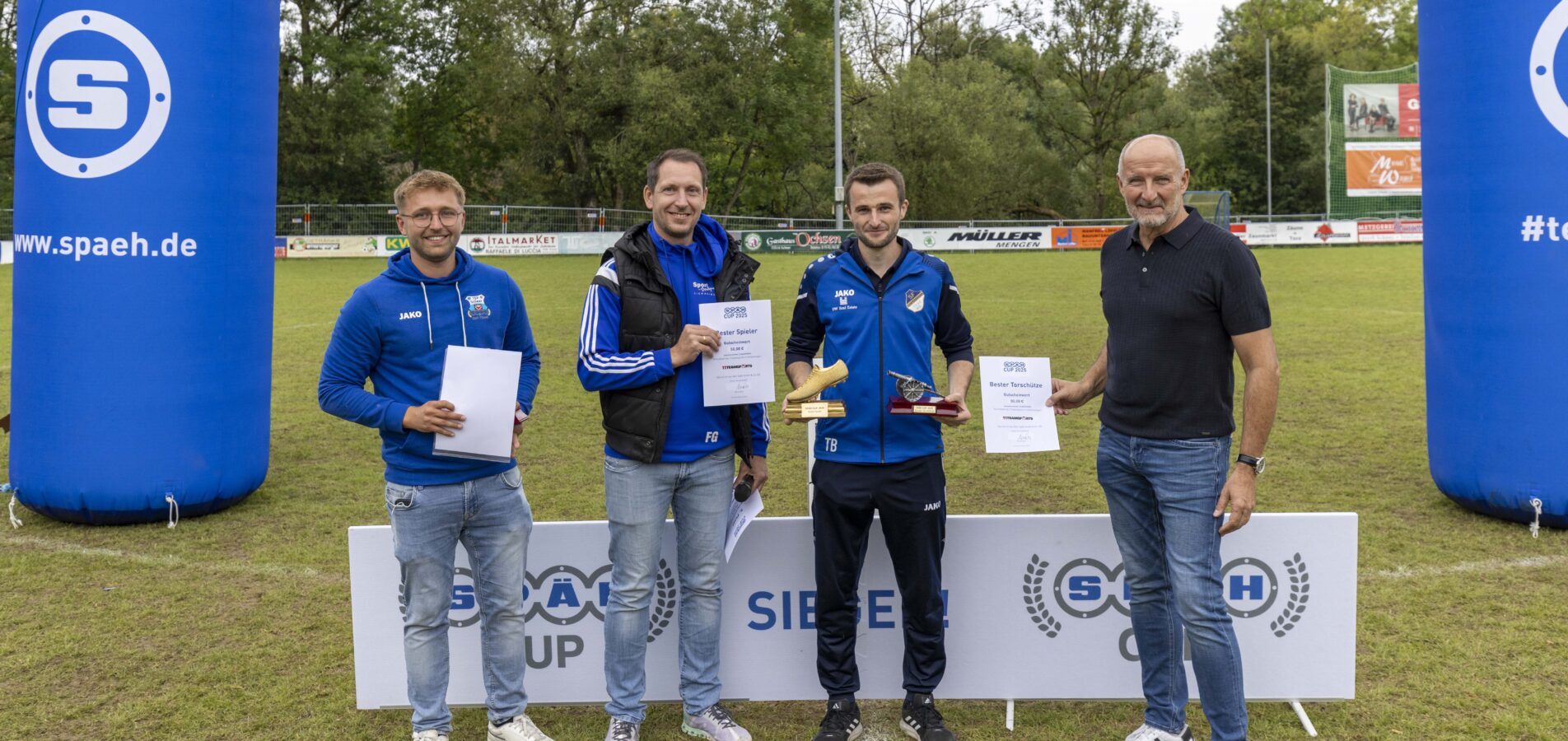 Vier Männer stehen auf einem Fußballfeld zwischen zwei großen, blauen SPÄH-Säulen und halten Urkunden und Pokale in die Kamera, wobei im Vordergrund eine Werbebande mit der Aufschrift SPÄH CUP SIEGER zu sehen ist.