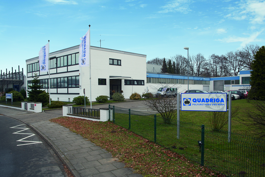 Das weiße, zweistöckige Bürogebäude der Firma Quadriga aus dem Jahr 1975 mit breiten Fensterfronten und zwei Fahnenmasten davor steht an einer Straße mit einem Zaun im Vordergrund.