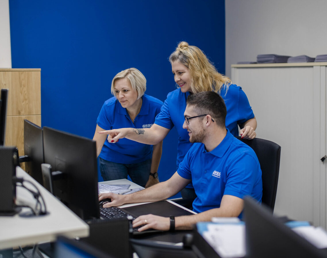 Ein Mitarbeiter der Späh-Gruppe sitzt am Schreibtisch und arbeitet am Computer, während zwei lächelnde Kolleginnen, eine davon zeigend, hinter ihm stehen und gemeinsam auf den Bildschirm schauen.