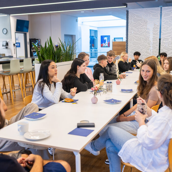 Alle Auszubildenden sitzen an einem Tisch in der Kantine um zu Frühstücken. Viele Lächeln dabei und unterhalten sich miteinander.