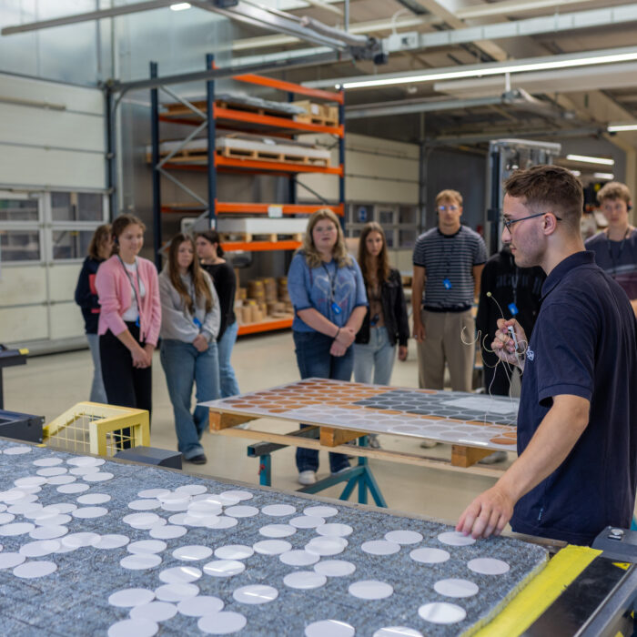 In der Produktion befindet sich eine Gruppe von Auszubildenden. Einer der Auszubildenden erklärt den anderen die Arbeitsprozesse und veranschaulicht diese anhand praktischer Beispiele.