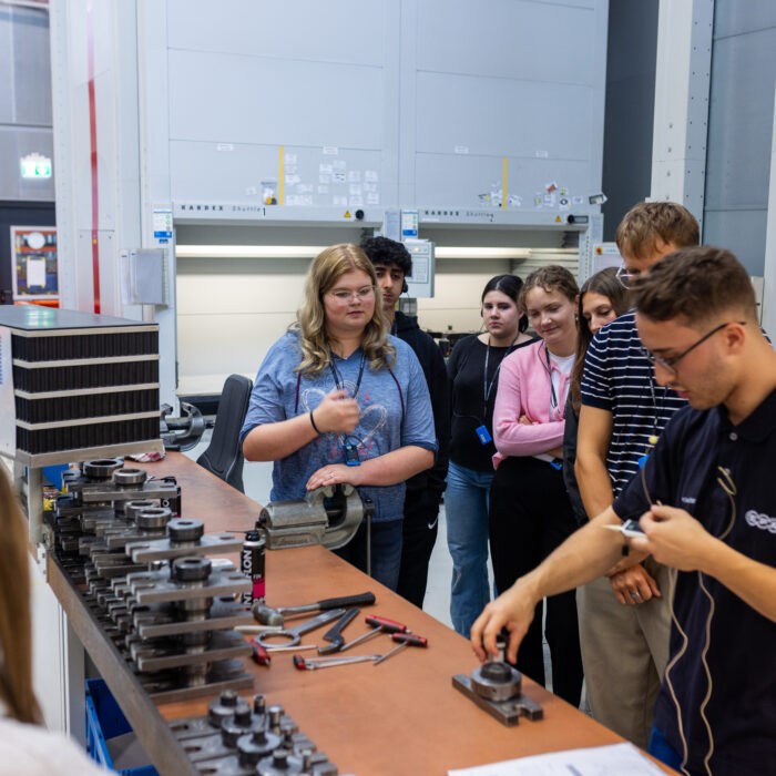 In der Produktion befindet sich eine Gruppe von Auszubildenden. Einer der Auszubildenden erklärt den anderen die Arbeitsprozesse und veranschaulicht diese anhand praktischer Beispiele.