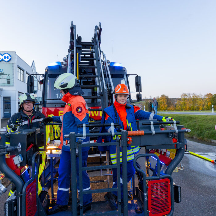 Zwei Mitarbeiter der Jugendfeuerwehr stehen im Korb der Drehleiter. Ein anderer Mitarbeiter der Feuerwehr steht daneben und hält einen Wasserschlauch. Das Feuerwehrfahrzeug steht vor dem Warenausgang.