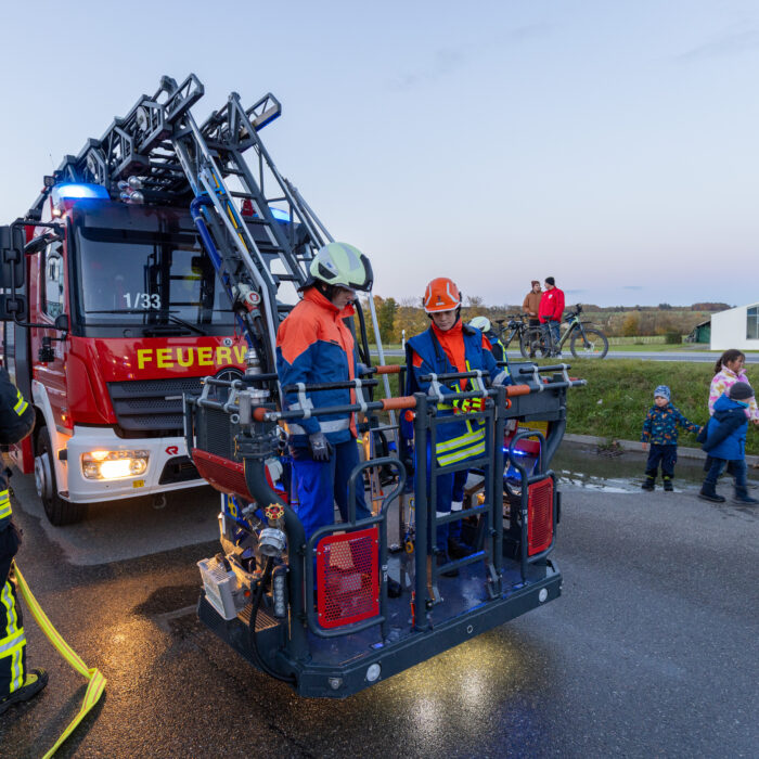 Im Korb der Drehleiter stehen zwei Mitarbeiter der Jugendfeuerwehr. Der ältere Mitarbeiter zeigt dem jüngeren Mitarbeiter wie der Korb geöffnet werden kann.