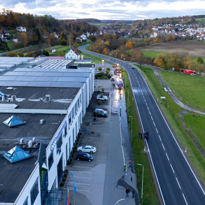 Zwei blinkende Feuerwehrfahrzeuge stehen vor dem Späh Gebäude. Zwei Mitarbeiter der Feuerwehr halten einen Wasserschlauch zusammen.