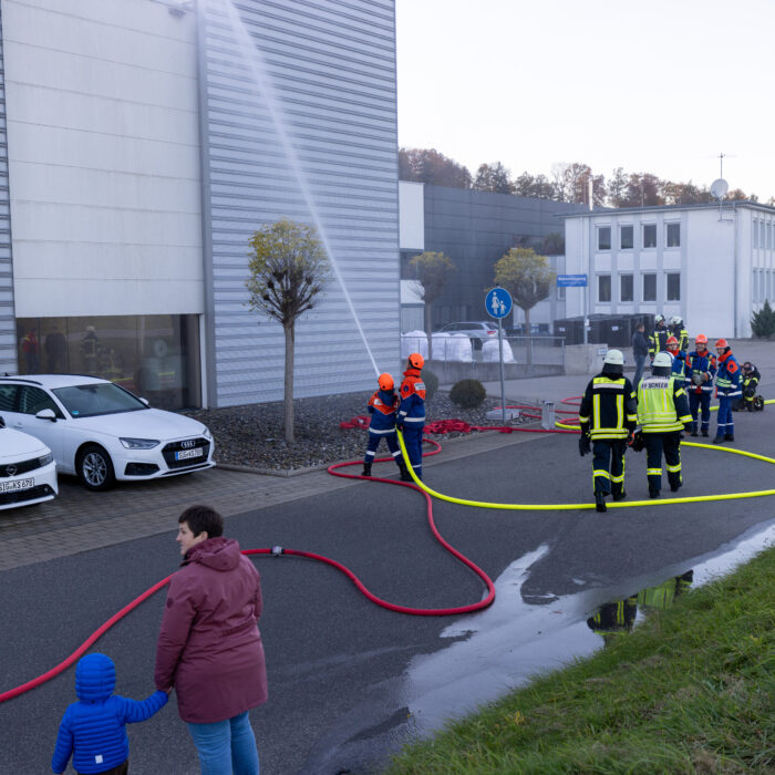 Zwei Kinder aus der Jugendfeuerwehr geben mit dem Schlauch ein Strahl auf das Späh Gebäude ab. Herum stehen weitere Menschen aus der Feuerwehr und Jugendfeuerwehr.