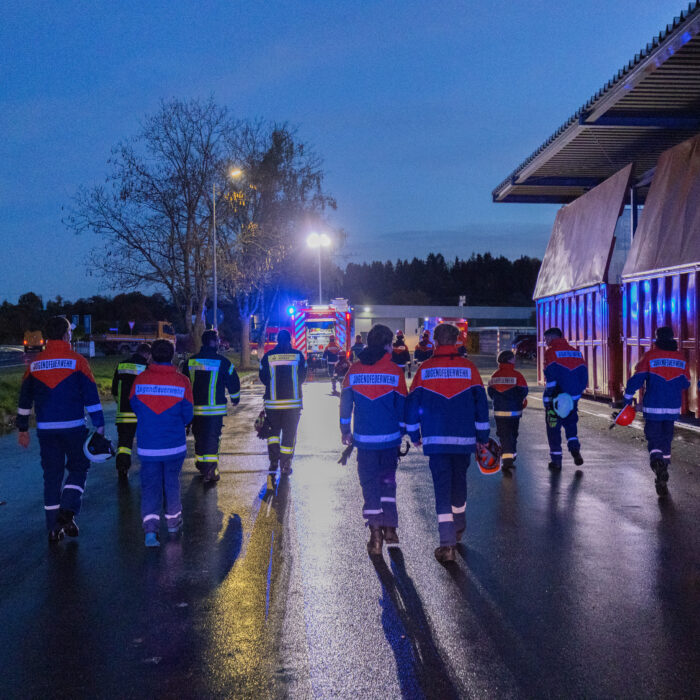 Mehrere Kinder aus der Jugendfeuerwehr und Erwachsene aus der Feuerwehr sind auf dem Späh Gelände für eine Übung. Sie gehen zusammen in die Richtung des blinkenden Feuerwehrfahrzeugs.