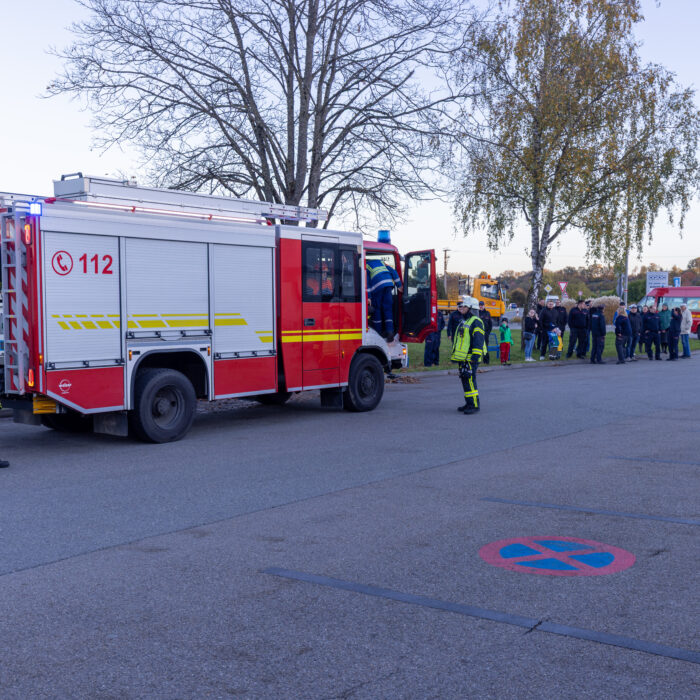 Ein Feuerwehrfahrzeug ist auf dem Späh Gelände. Davor stehen drei Menschen aus der Feuerwehr. Im Hintergrund sind Zuschauer die in Richtung des Fahrzeugs schauen.