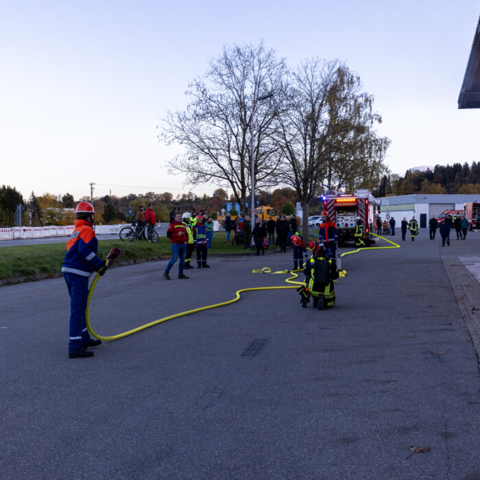 Ein Junge aus der Jugendfeuerwehr hält den langen Feuerwehrschlauch. Ein Mann aus der Feuerwehr und 3 andere Kinder aus der Jugendfeuerwehr machen Vorbereitungen. Im Hintergrund stehen mehrere Menschen die zuschauen.