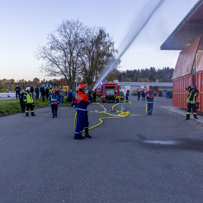 Zwei Kinder aus der Jugendfeuerwehr üben mit dem Schlauch ein Strahl abzugeben auf die Überdachung der Müllcontainer. Herum steht ein Mann aus der Feuerwehr der ein Bild schießt und weitere Zuschauer.