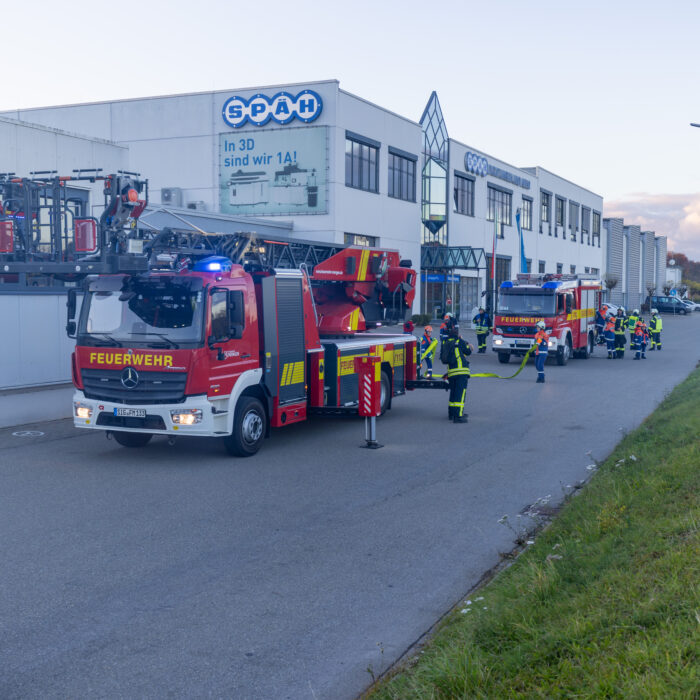 Vor dem Späh Gebäude stehen zwei Feuerwehrfahrzeuge. Um die Fahrzeuge herum stehen beschäftigte Kinder aus der Jugendfeuerwehr und Erwachsene aus der Feuerwehr.