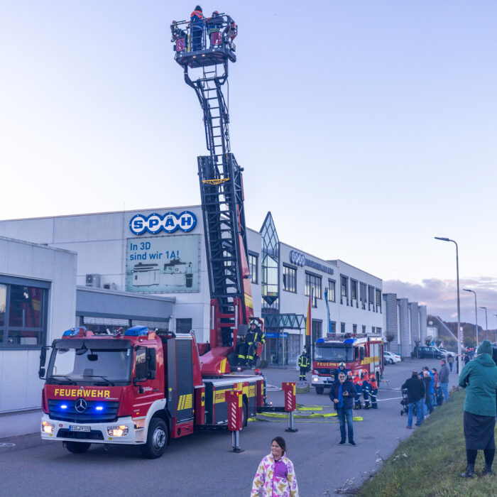 Vor dem Warenausgang steht ein Feuerwehrfahrzeug. Ein Mann aus der Feuerwehr fährt die Drehleiter nach oben. Im Korb der Drehleiter sind zwei Kinder aus der Jugendfeuerwehr. Mehrere Menschen schauen dabei zu.