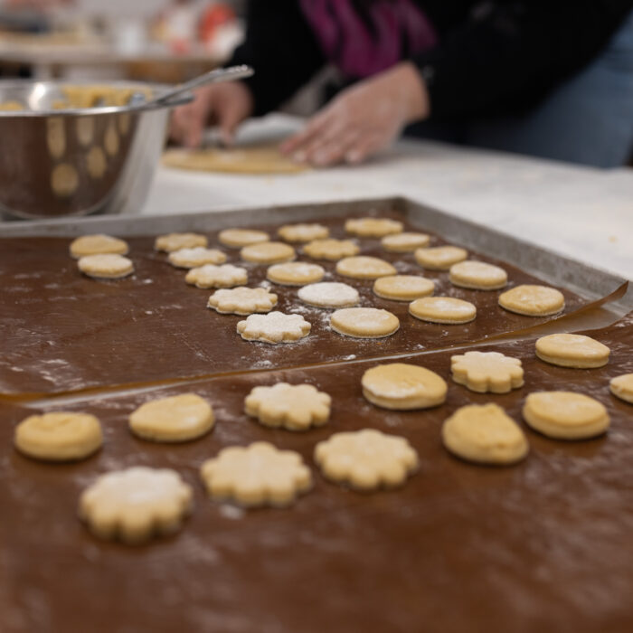 Weihnachtsgebäck ist in verschiedensten Formen auf dem Backblech. Im Hintergrund ist eine Schüssel mit Teig und ein Späh-Mitarbeiter der Plätzchen backt.