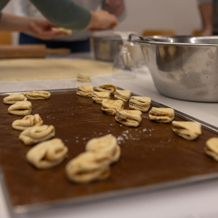In der Kantine ist ein Backblech mit Weihnachtsgebäck und Schüsseln. Im Hintergrund sind zwei Späh-Mitarbeiter mit einem ausgerollten Teig.