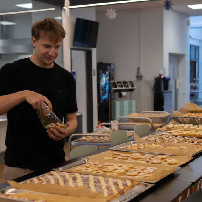 Auf einem langen Tisch stehen viele Bleche mit unterschiedlichen fertigen Weihnachtsgebäcken. Ein Späh-Mitarbeiter steht vor dem Tisch und hält eine Packung in der Hand.