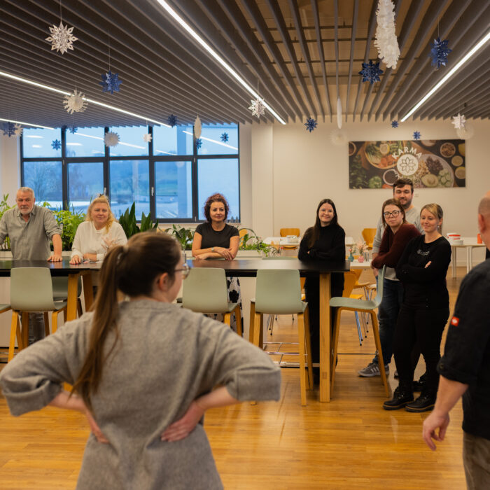 Viele Späh-Mitarbeiter und ein Koch stehen versammelt in der Kantine. Der Koch erklärt den Späh-Mitarbeitern das Vorgehen für das Backen vom Weihnachtsgebäck.