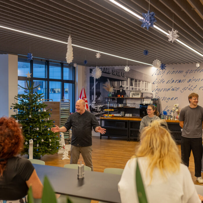 Viele Späh-Mitarbeiter und der Koch stehen versammelt in der Kantine. Der Koch ist am lächeln und erklärt den Mitarbeitern etwas mit offenen Armen.